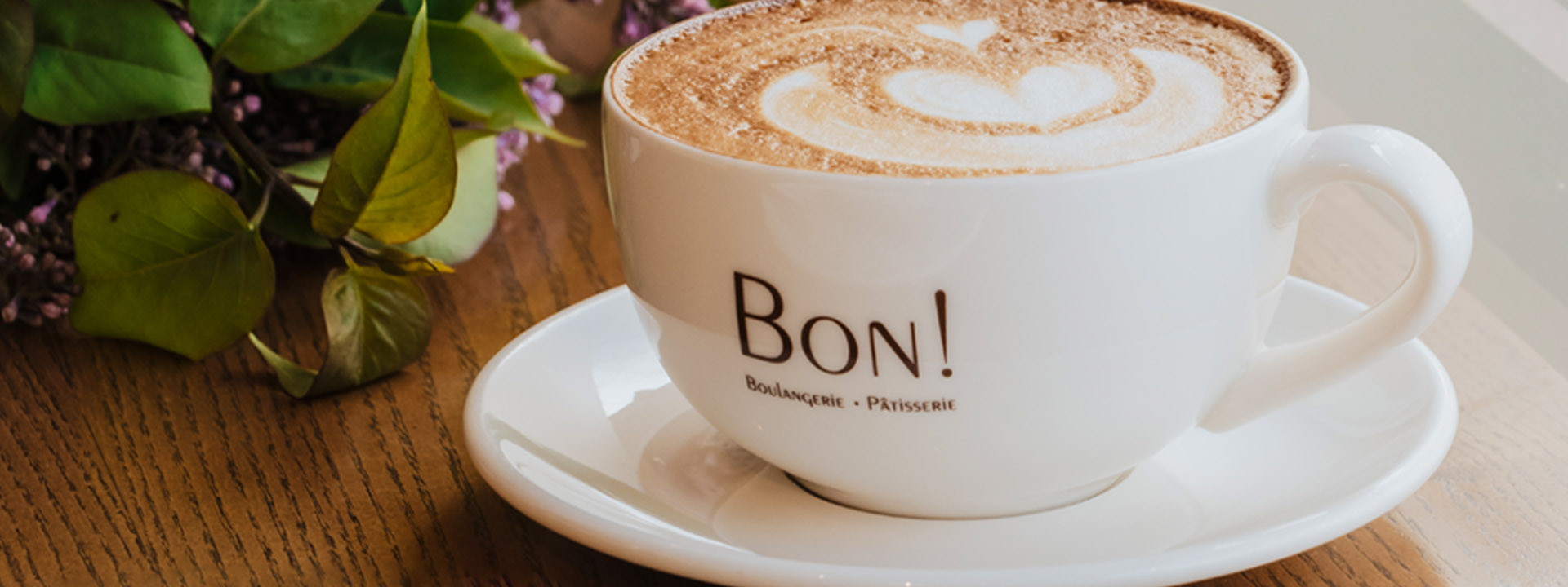 A cup of cappuccino with latte art on a saucer, featuring the text "BON! Boulangerie - Pâtisserie" on the cup. The cup is placed on a wooden table next to some green leaves.