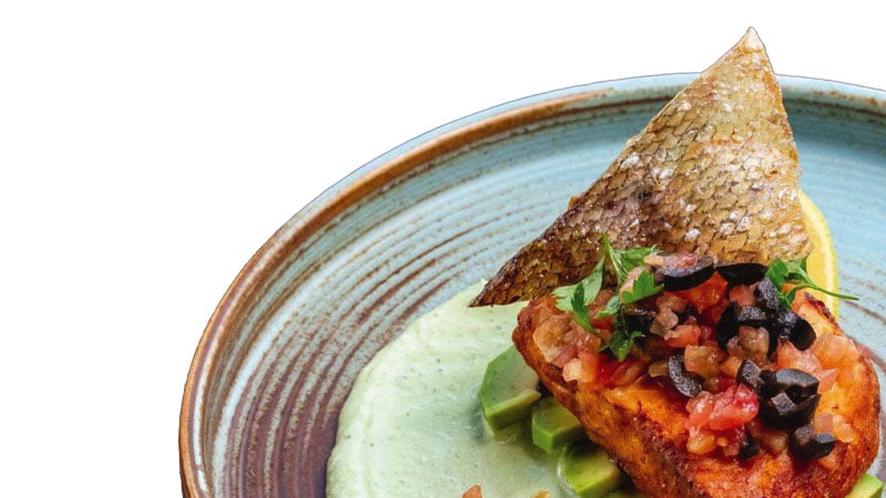 A plated dish featuring a piece of cooked fish with crispy skin, topped with diced tomatoes and olives, served on a green sauce.