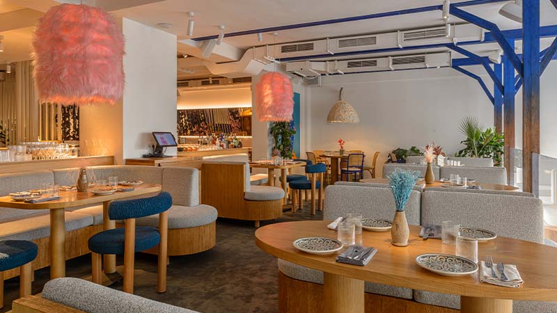 Modern restaurant interior with wooden tables, cushioned seating, pink and wicker pendant lights, and table settings, featuring a mix of natural and contemporary decor elements.