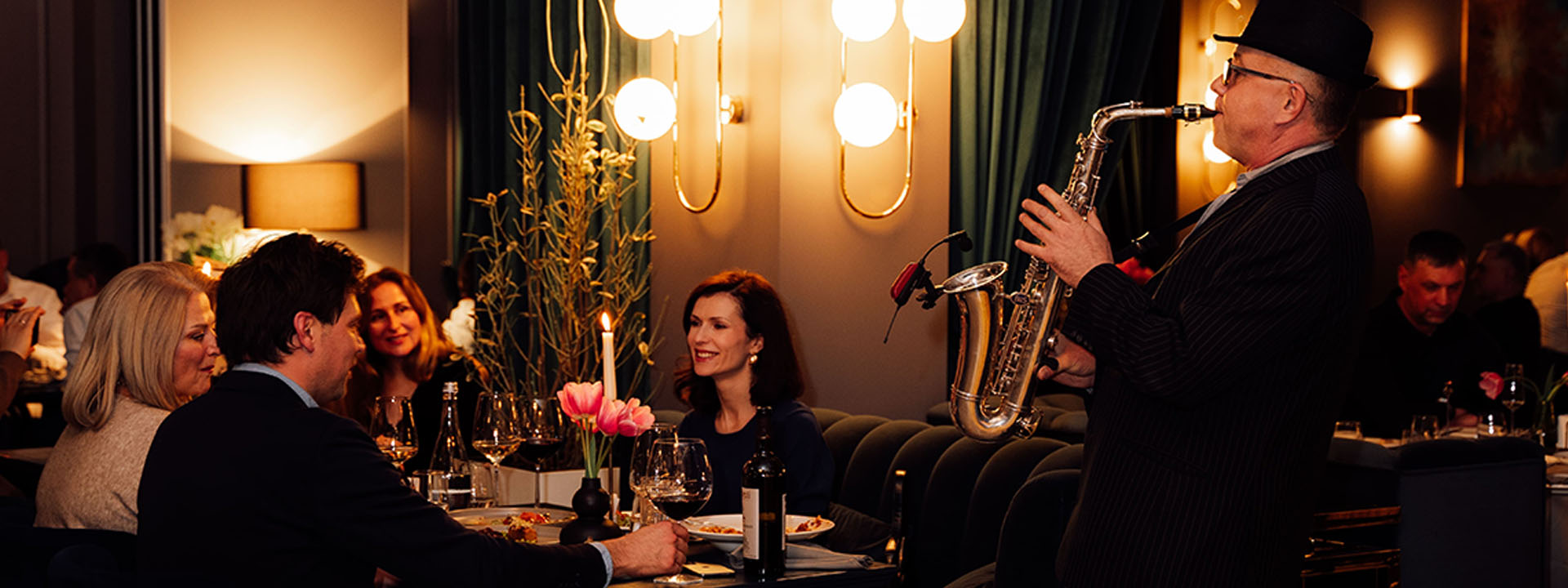 A group of people dining at a restaurant while a man in a hat and suit plays the saxophone next to them.