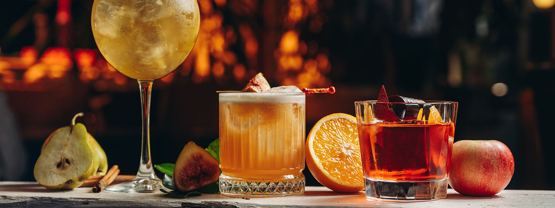 Three cocktails on a bar counter with fresh fruits including pear, fig, orange, and apple in a warm, dimly-lit setting.