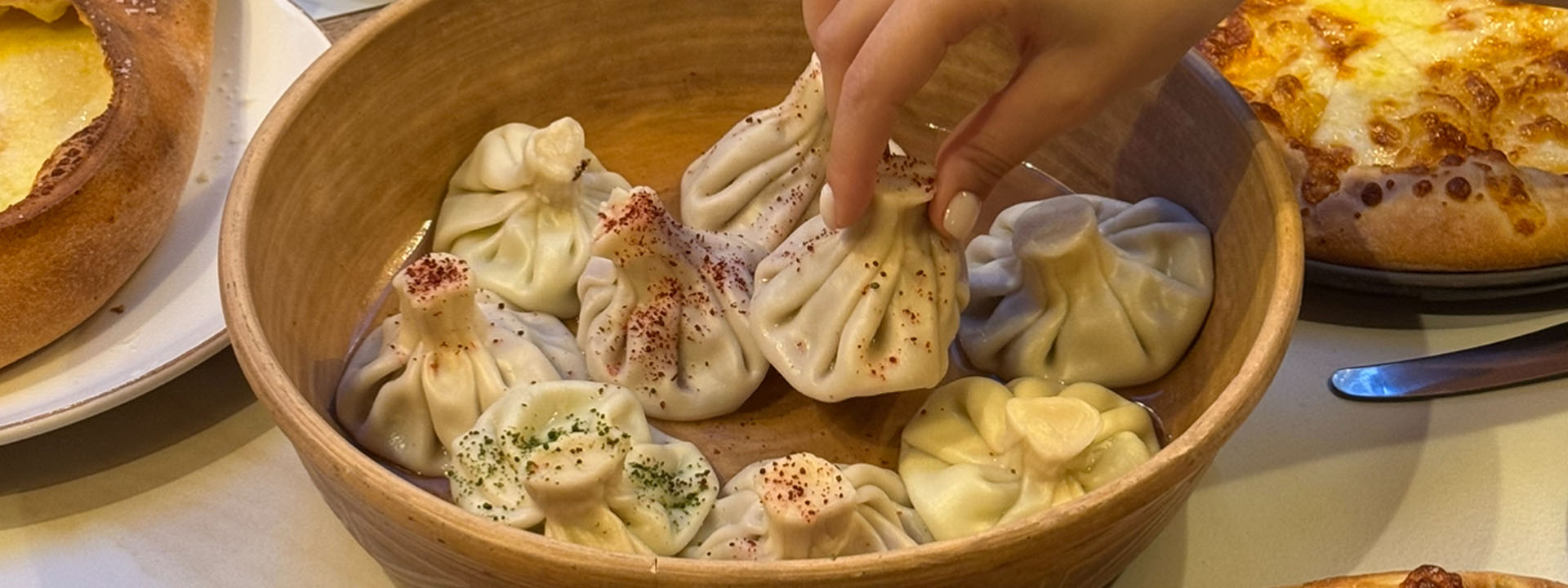 A hand picks up a dumpling from a wooden bowl filled with khinkali, surrounded by other dishes on a table.