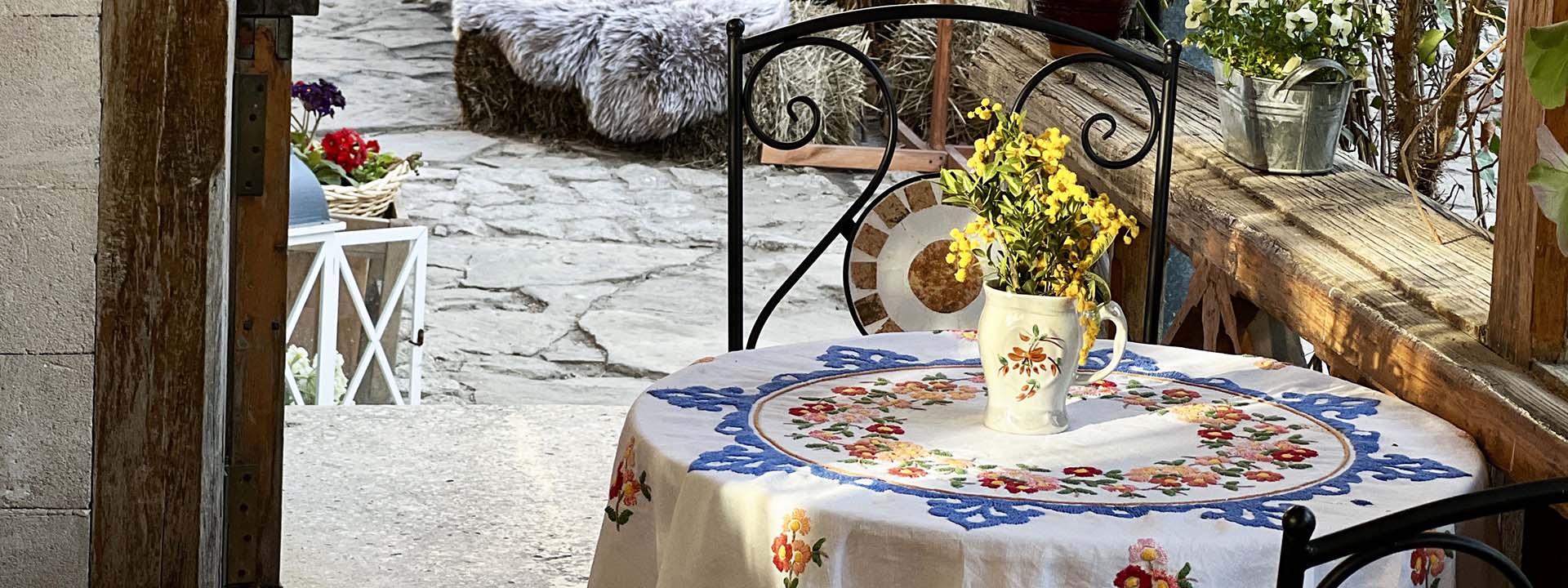 A round table with a floral embroidered tablecloth and a jug of yellow flowers sits by a window, with a stone patio and various potted plants visible outside.