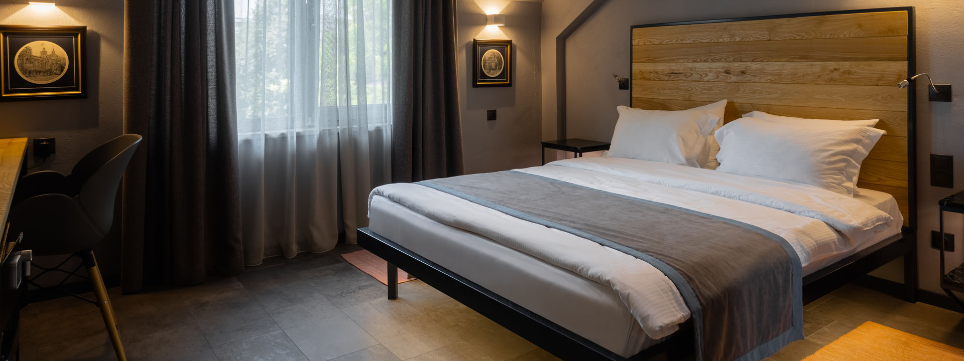 Modern hotel room with a large bed, wooden headboard, two framed pictures on the wall, a window with sheer curtains, and a desk with a chair in the corner.