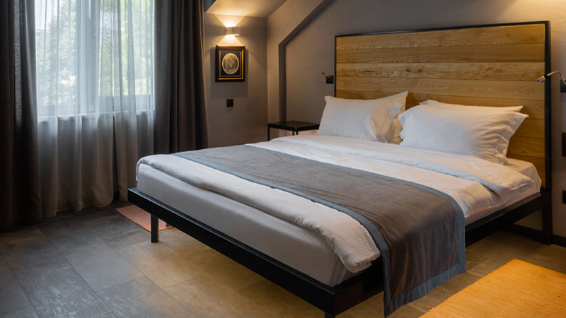 Modern hotel room with a large bed, wooden headboard, two framed pictures on the wall, a window with sheer curtains, and a desk with a chair in the corner.