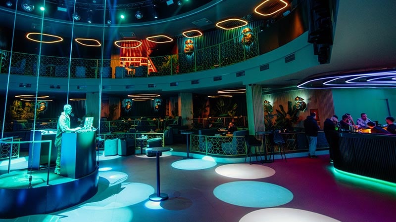 A DJ stands at a booth in the center of a modern, colorful nightclub with circular seating, neon lights, and a few people at the bar.