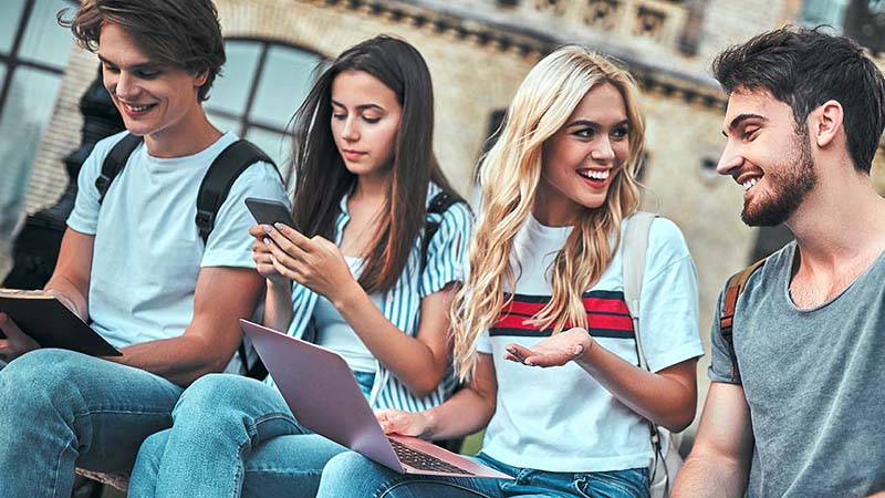 Four students sitting outdoors, with one reading a book, another using a smartphone, a third looking at a laptop, and all casually interacting.