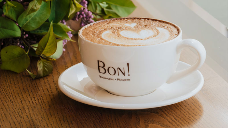 A cup of cappuccino with latte art on a saucer, featuring the text "BON! Boulangerie - Pâtisserie" on the cup. The cup is placed on a wooden table next to some green leaves.
