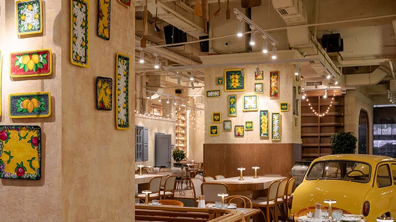 A brightly lit restaurant interior with framed art on the walls, wooden tables and chairs, and part of a yellow car displayed inside.