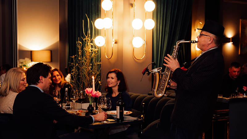 A group of people dining at a restaurant while a man in a hat and suit plays the saxophone next to them.