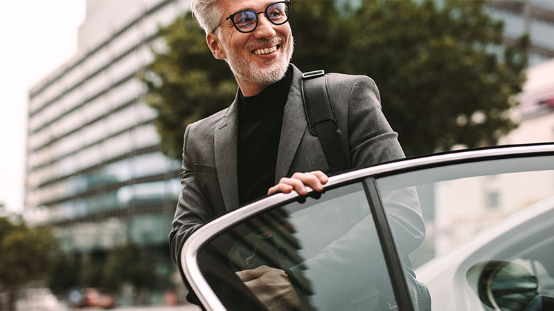 A man in a suit and glasses smiles while closing the door of a car in an urban area.