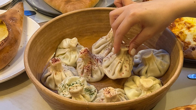 A hand picks up a dumpling from a wooden bowl filled with khinkali, surrounded by other dishes on a table.