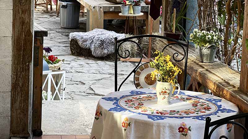 A round table with a floral embroidered tablecloth and a jug of yellow flowers sits by a window, with a stone patio and various potted plants visible outside.