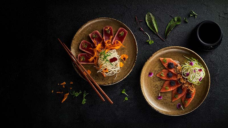 Two ceramic plates with artfully arranged tuna sashimi, noodles, garnishes, and edible flowers on a dark surface; chopsticks and a small cup are also visible.