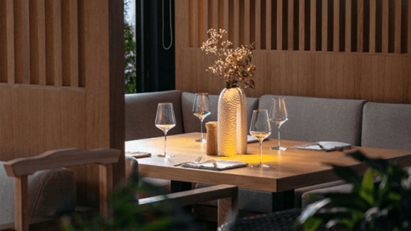 A wooden dining table set for four with wine glasses, cloth napkins, and a decorative vase with dried flowers in a modern restaurant setting.