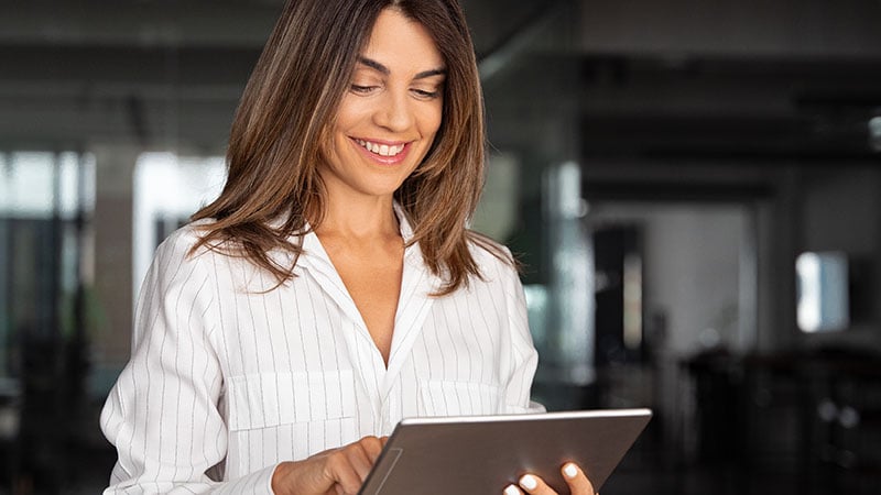 A woman wearing a white blouse is smiling and looking at a tablet in a modern office setting.